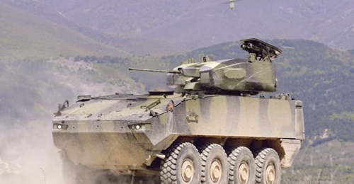 A Piranha 5 8x8 fitted with Rafael's Samson Integrated RWS armed with a 30 mm cannon, pod of two LR2 ATGWs, and a Spike Firefly UAV hovering above.